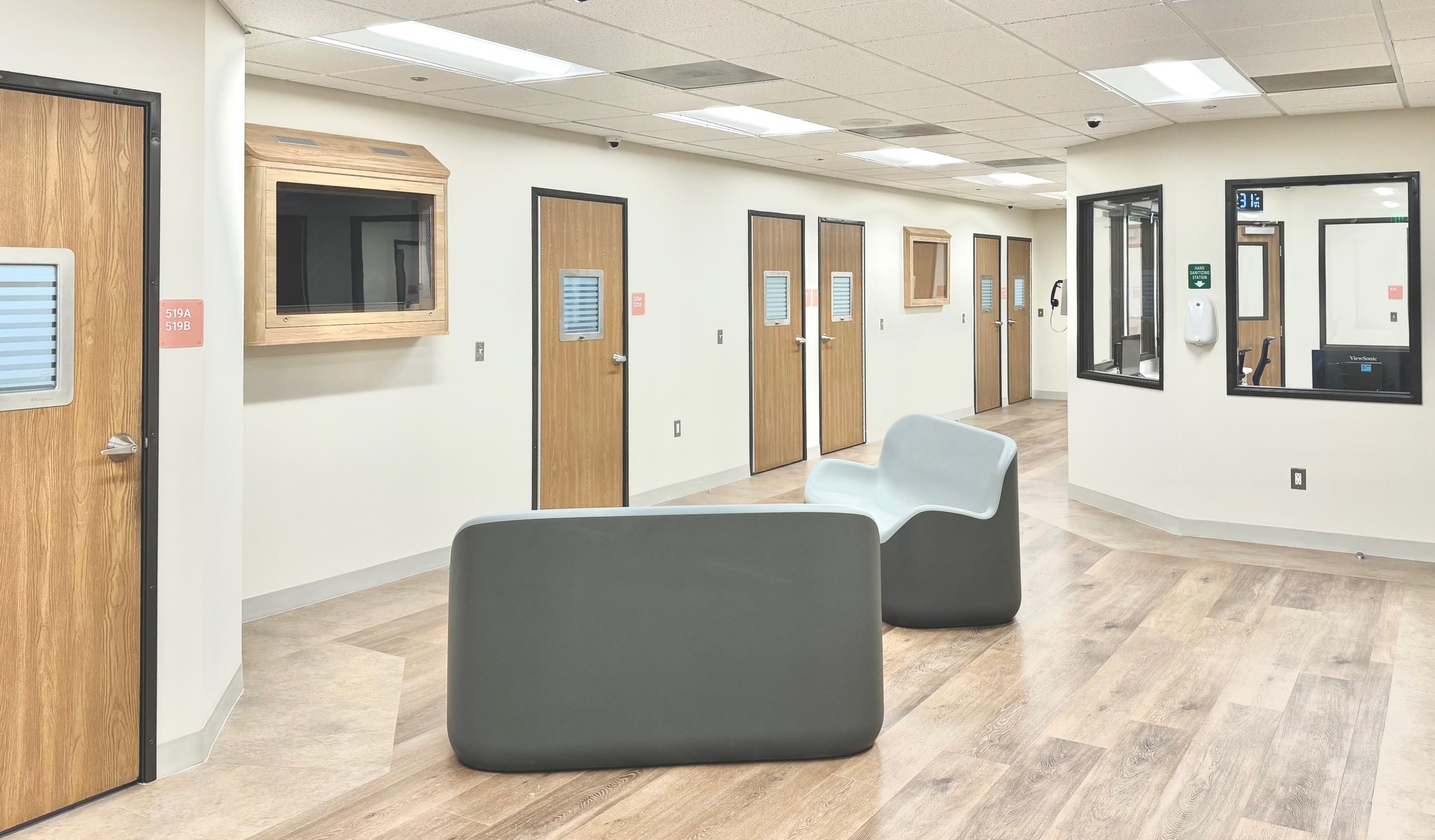 Modern healthcare facility hallway with wood doors featuring small windows, secure seating in the center, and a reception area with a glass window and wall-mounted sanitizer dispenser.