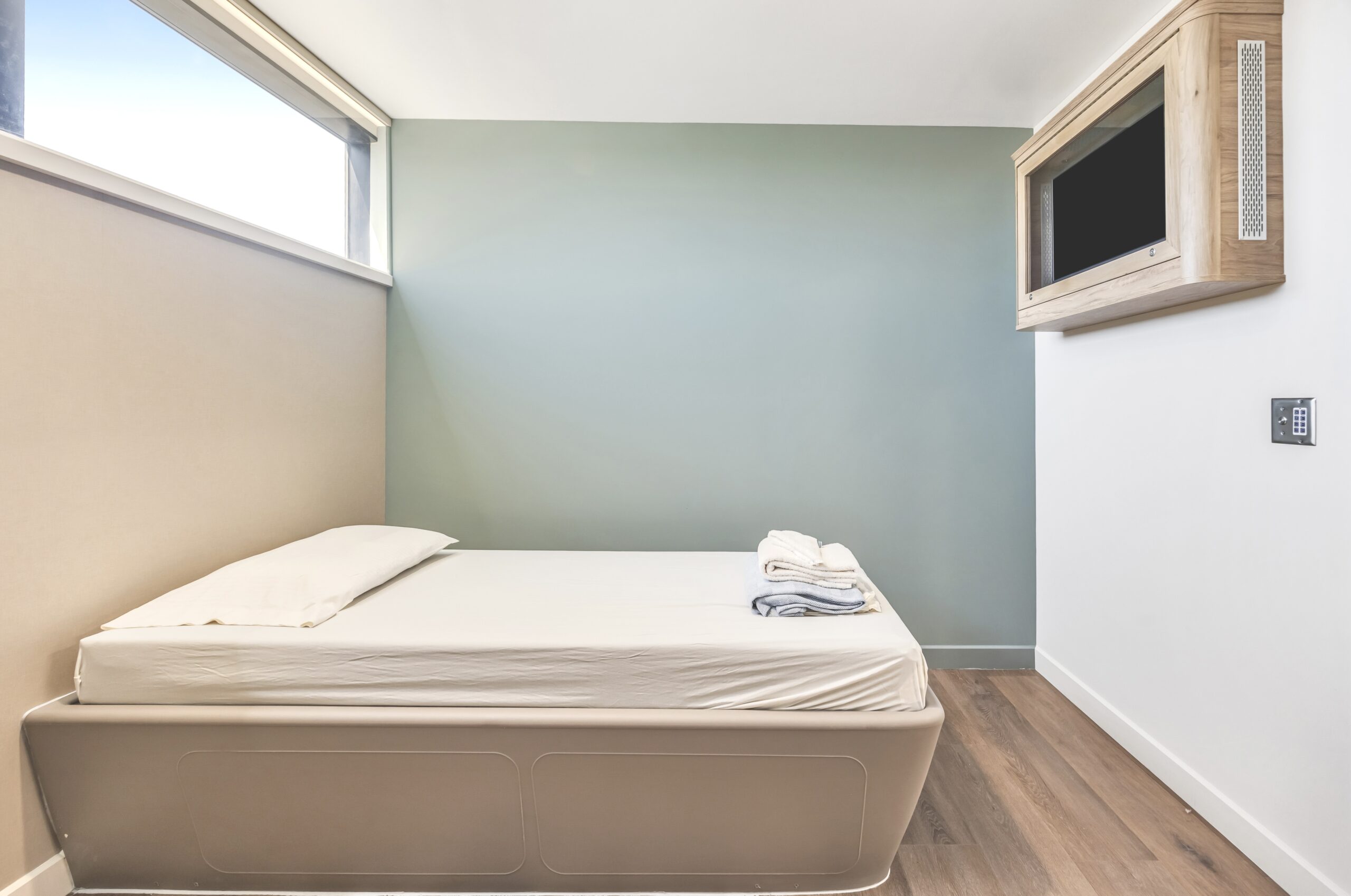 Minimalist patient room with single bed, folded linens, mounted television in wooden frame, and high window providing natural light.