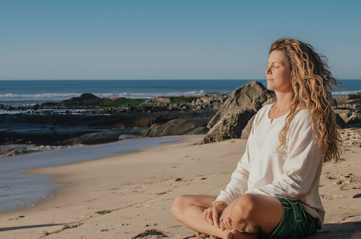 Woman sitting cross-legged on the beach meditating peacefully with eyes closed, facing the ocean under clear blue sky.