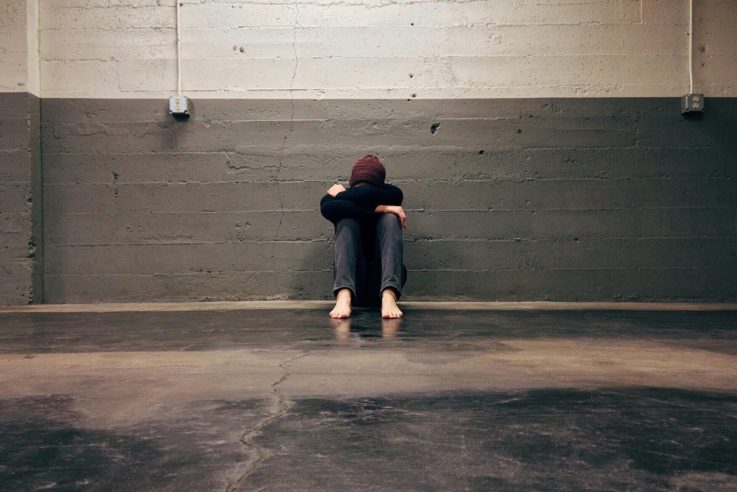 Person sitting barefoot on the floor against a concrete wall, hugging their knees and resting their head on their arms, conveying sadness or isolation.