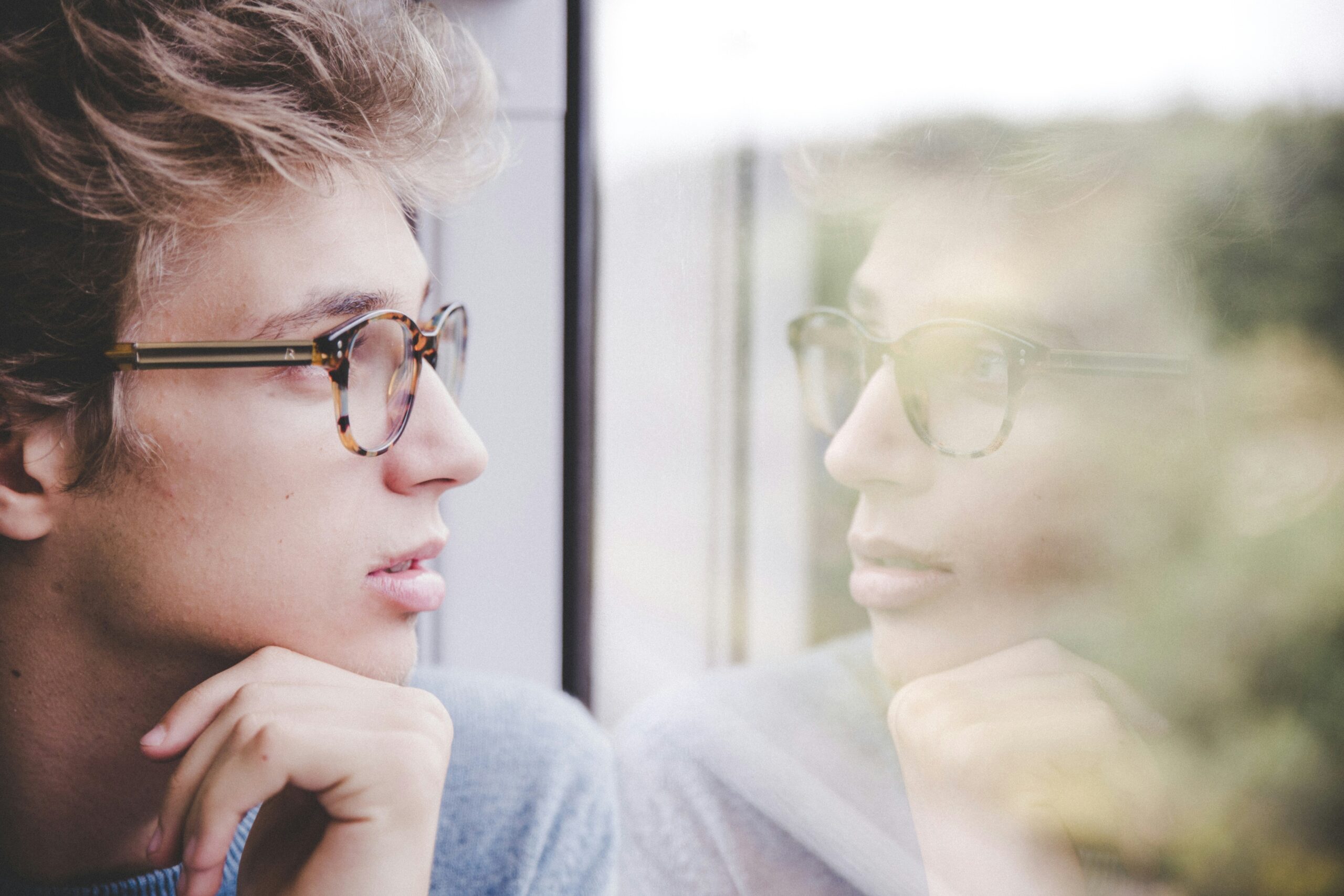 Young person wearing glasses looking out a window in deep thought